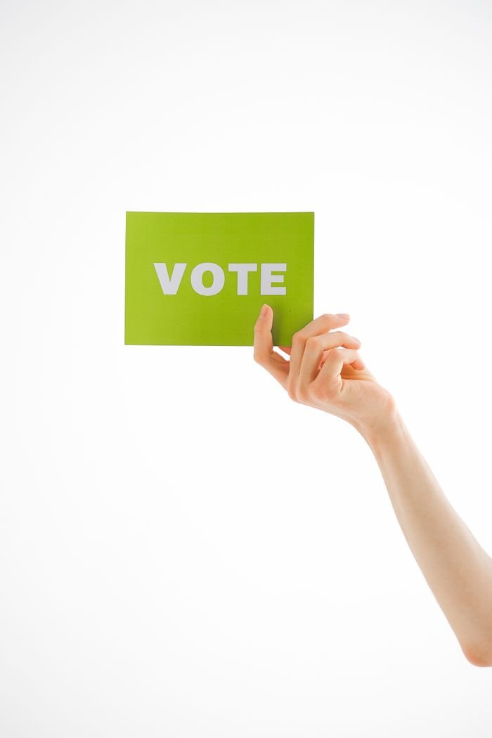 A hand raising a green card with the word 'VOTE' on a white background, offering ample copyspace.